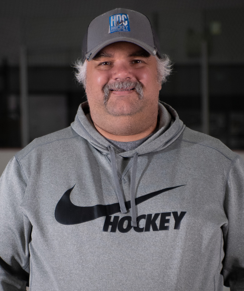Smiling person in gray hoodie and cap with hockey logo, standing indoors.