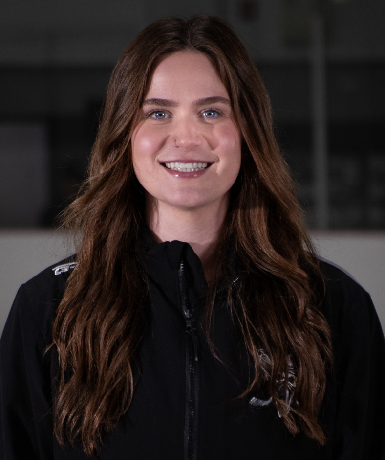 Smiling woman with long brown hair wearing a black zip-up jacket against a dark indoor background