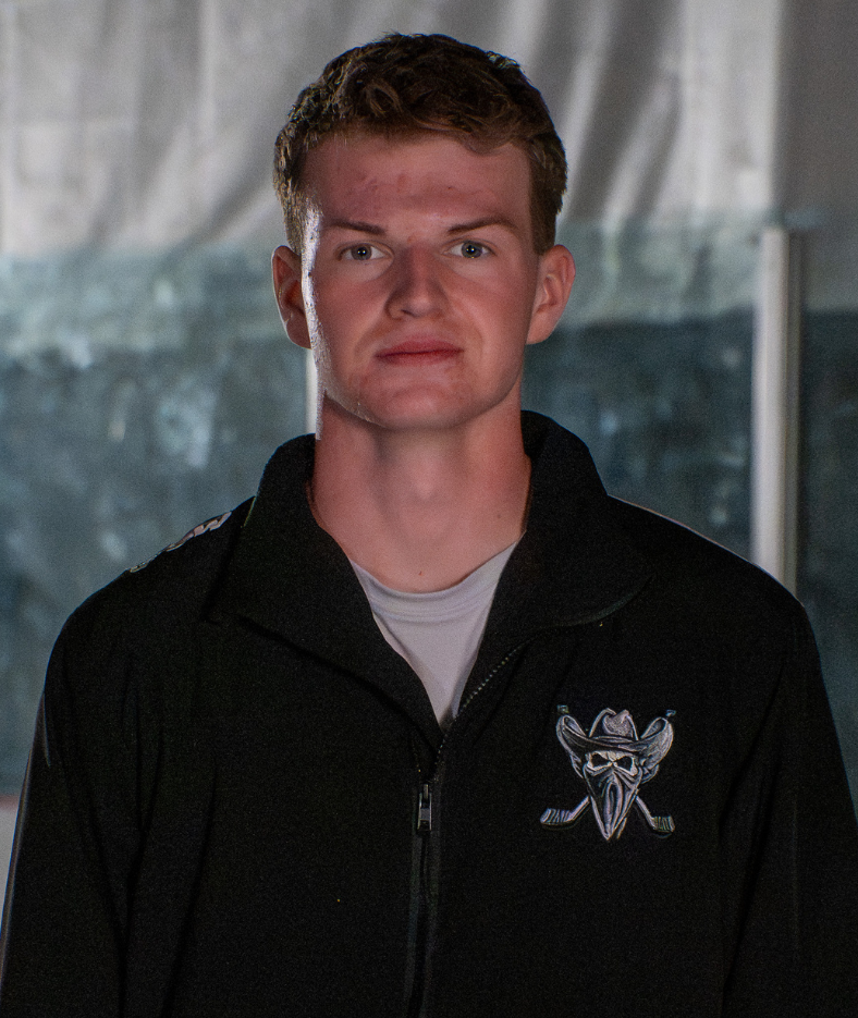 Portrait of a man in a black jacket with a winged emblem, standing indoors against a gray background