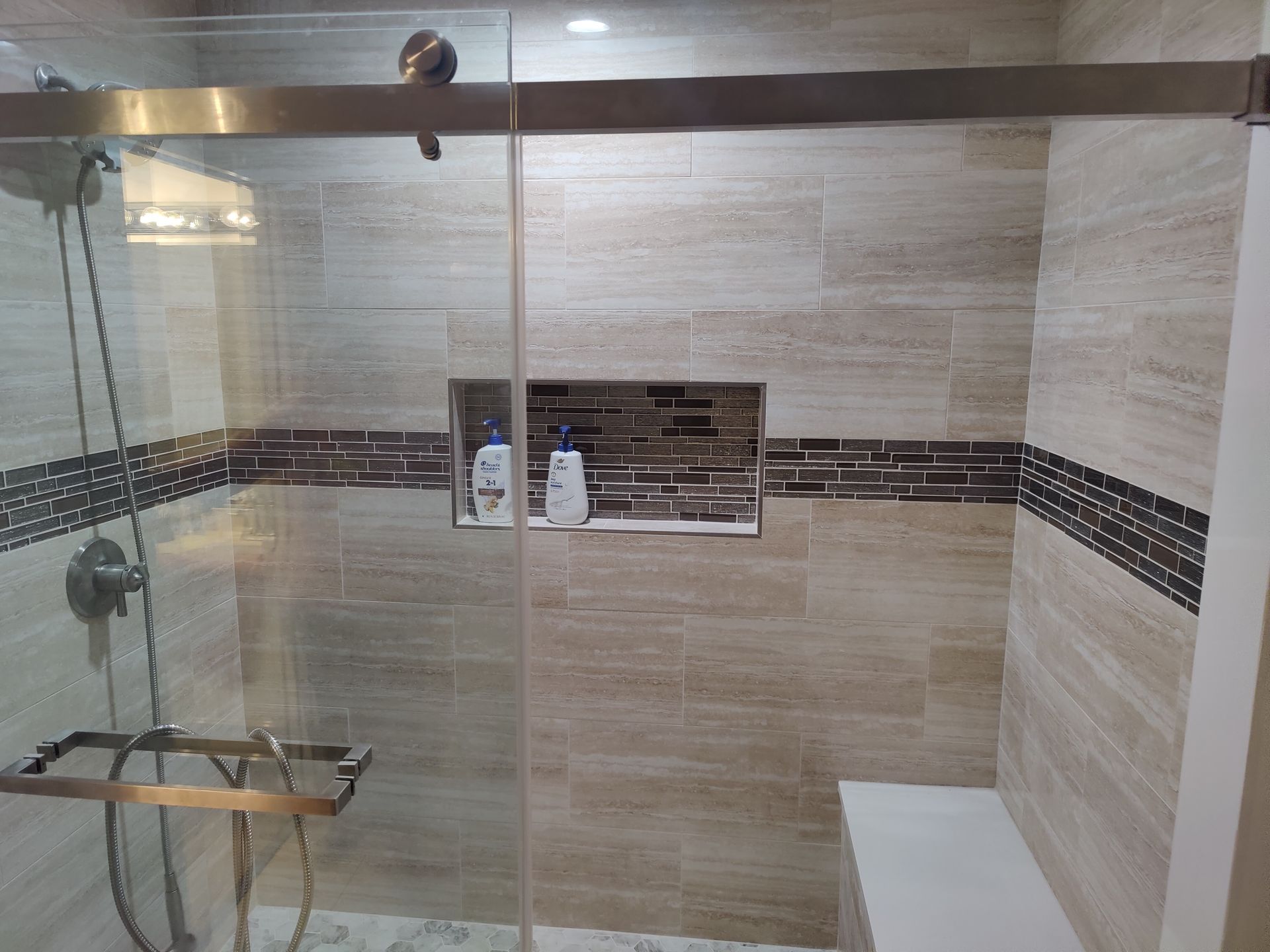 Modern bathroom shower with glass door, beige tiled walls, dark accent strip, and built-in bench