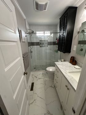 Small white bathroom with glass shower, marble-pattern floor, vanity, and window with black curtains.