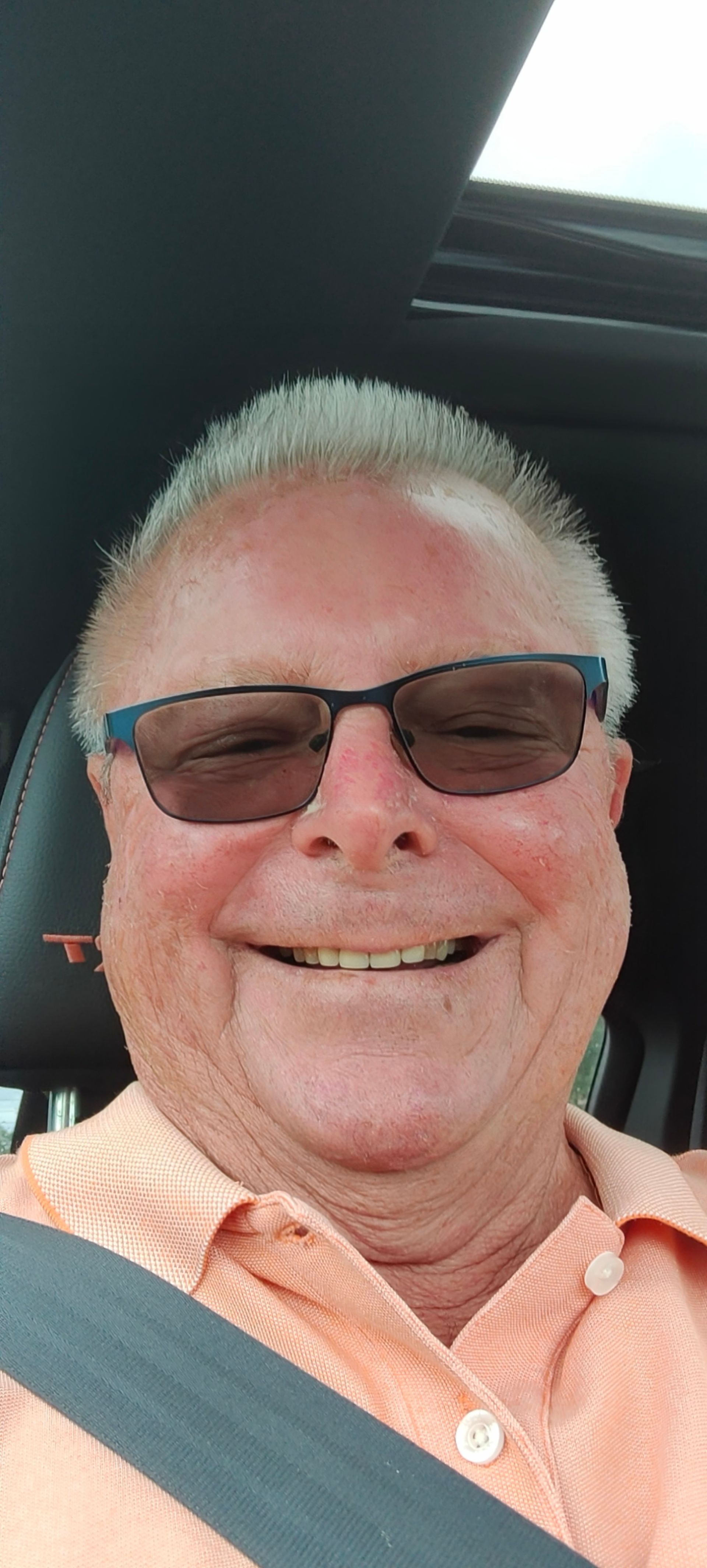 Smiling person wearing sunglasses and a peach shirt in a car, with a seat belt on.