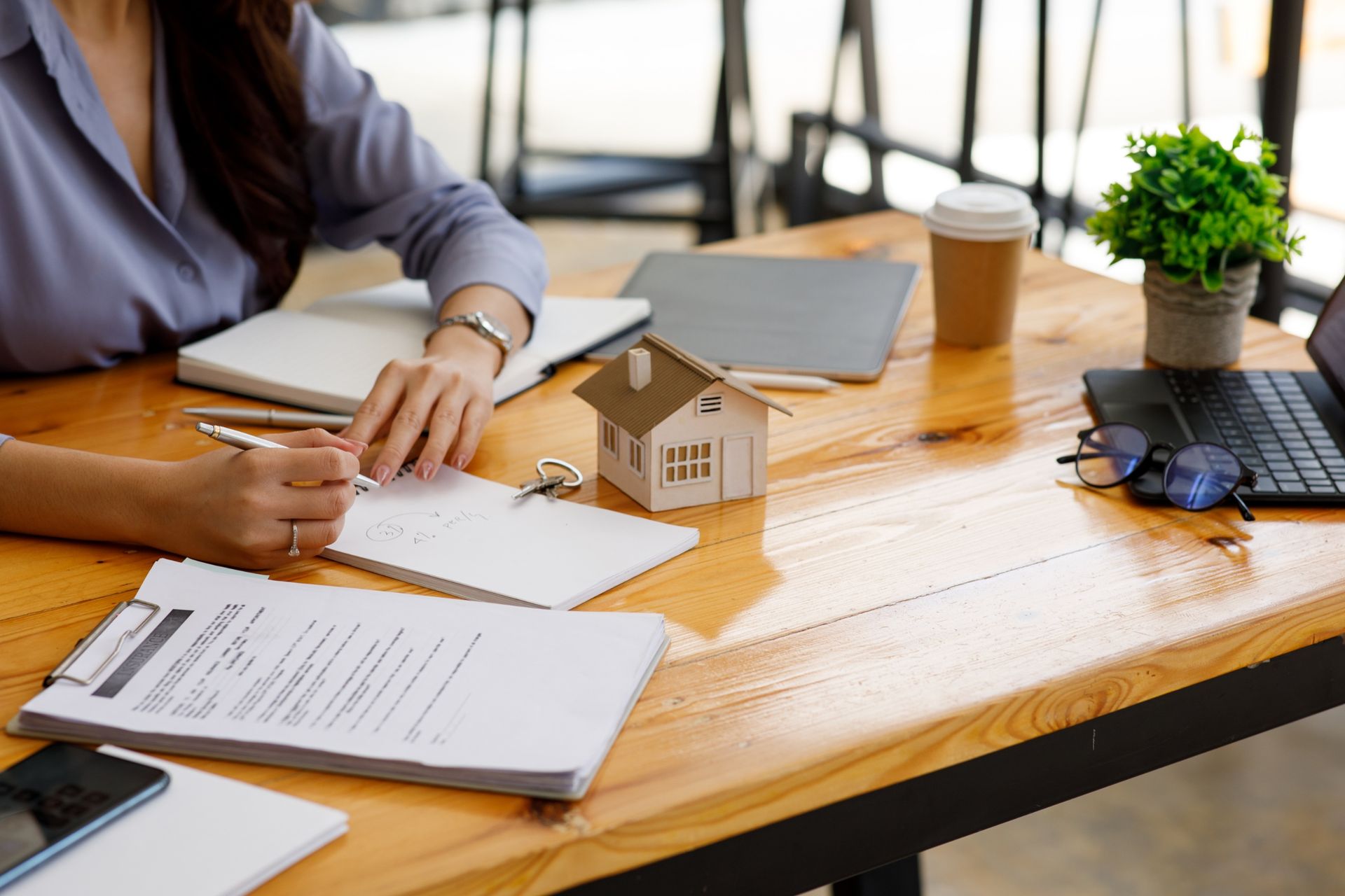 Person reviewing documents at a wooden desk with a house model, laptop, coffee cup, and plant