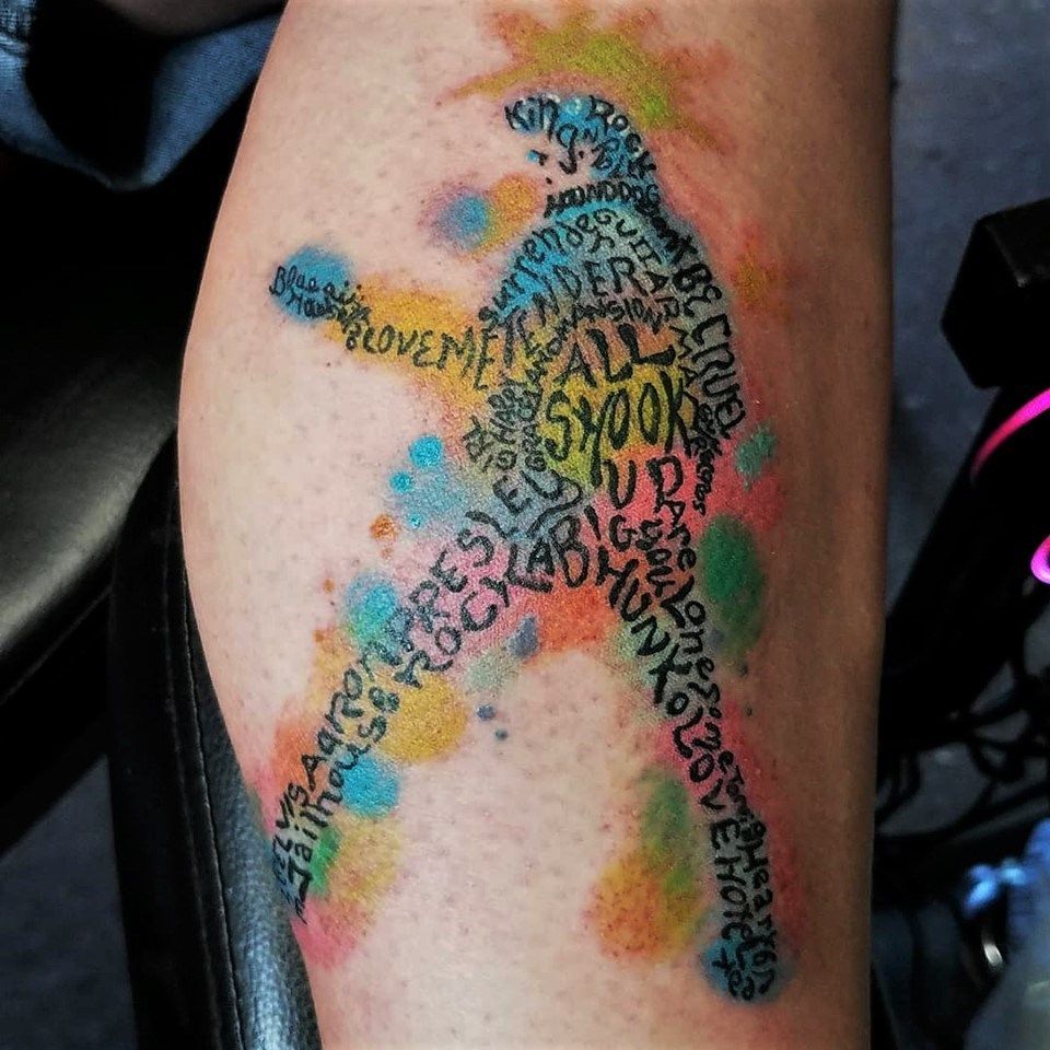 Tattoo of Elvis Presley silhouette filled with song titles, against colorful watercolor splashes on skin.