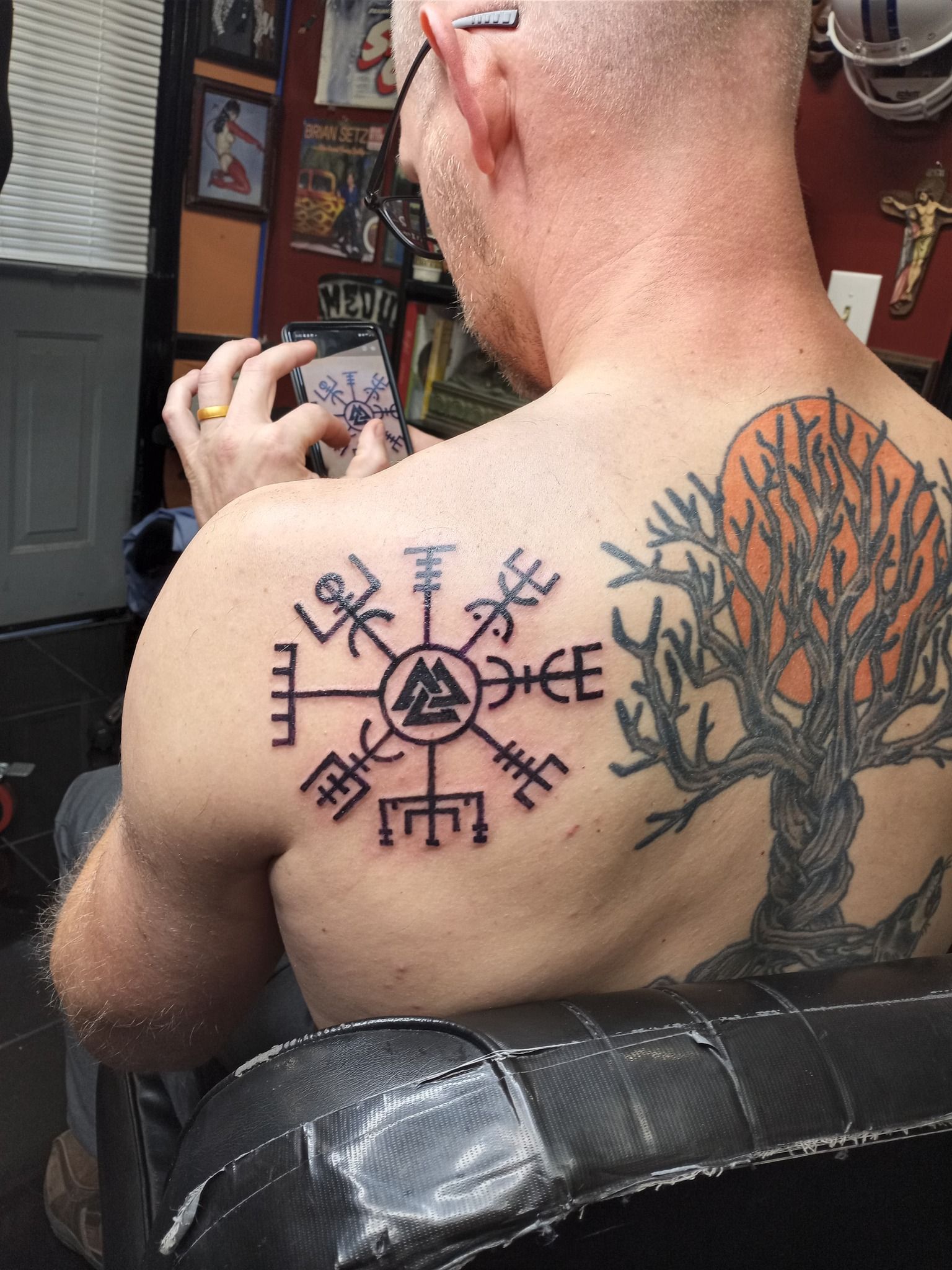 Man with two back tattoos sits holding a phone. Large tattoos include a compass-like symbol and a tree.