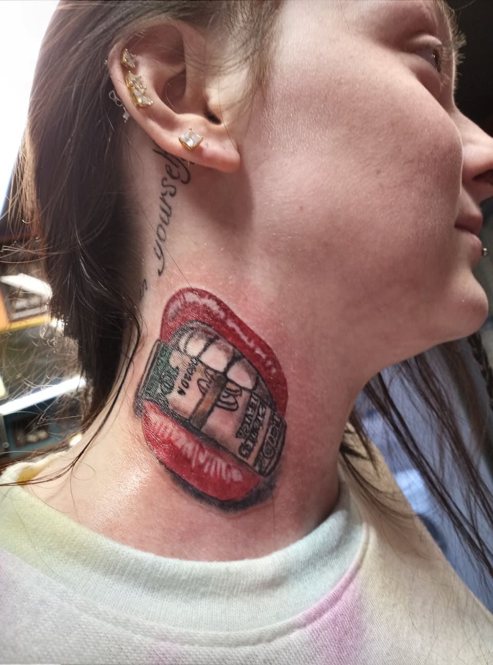 Woman's neck tattoo: red lips biting a wad of cash. She is wearing a white top.