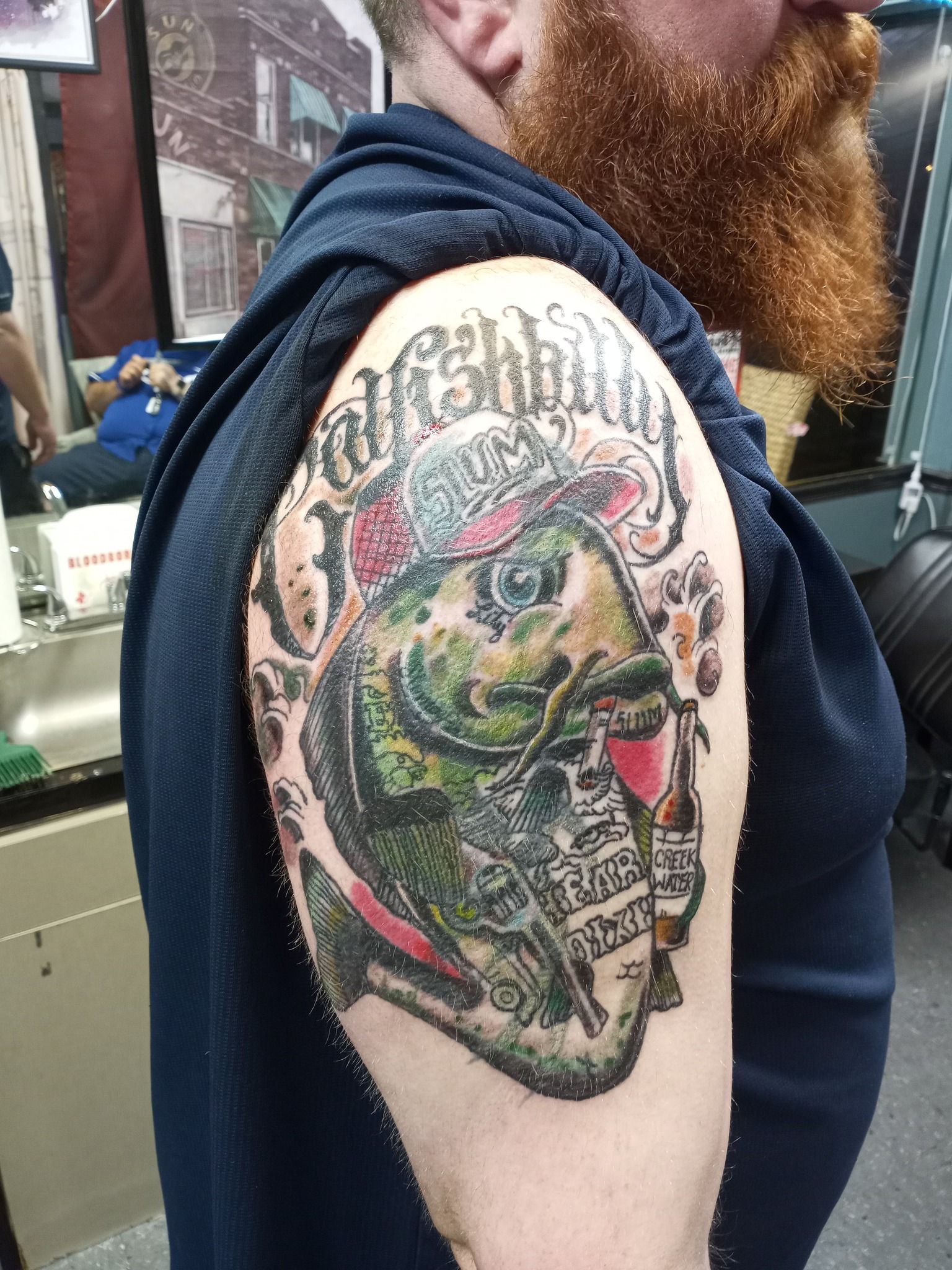 Tattoo of a fish with a hat and beer can on a man's upper arm.