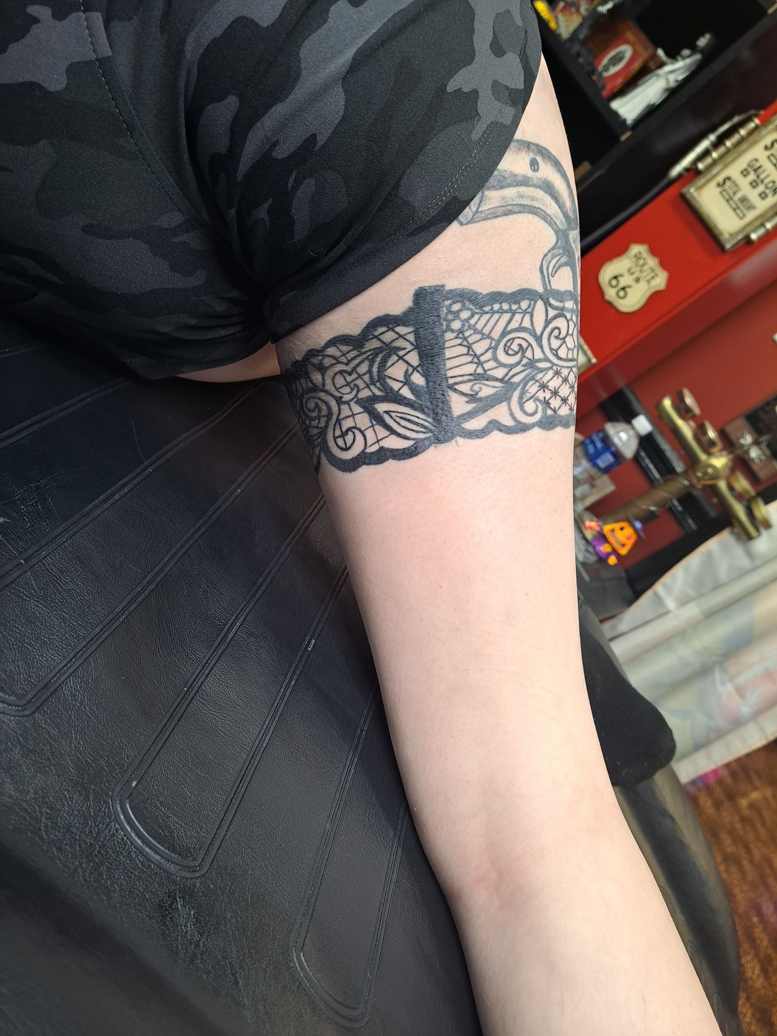 Arm with a black lace band tattoo; person in black top; tattoo parlor background.