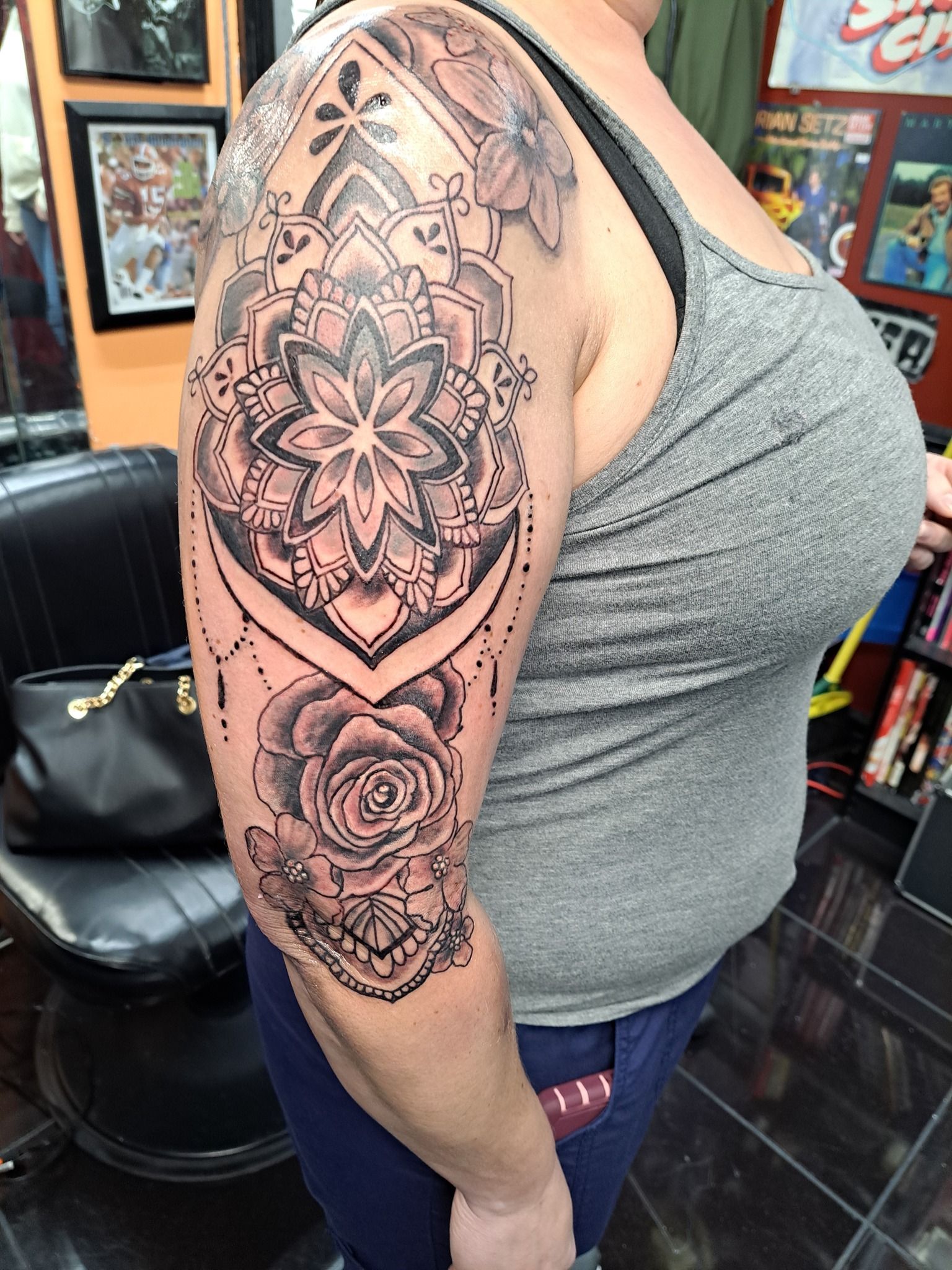 Woman with a gray tank top, showing off a large black mandala and rose tattoo on her arm. Tattoo shop setting.