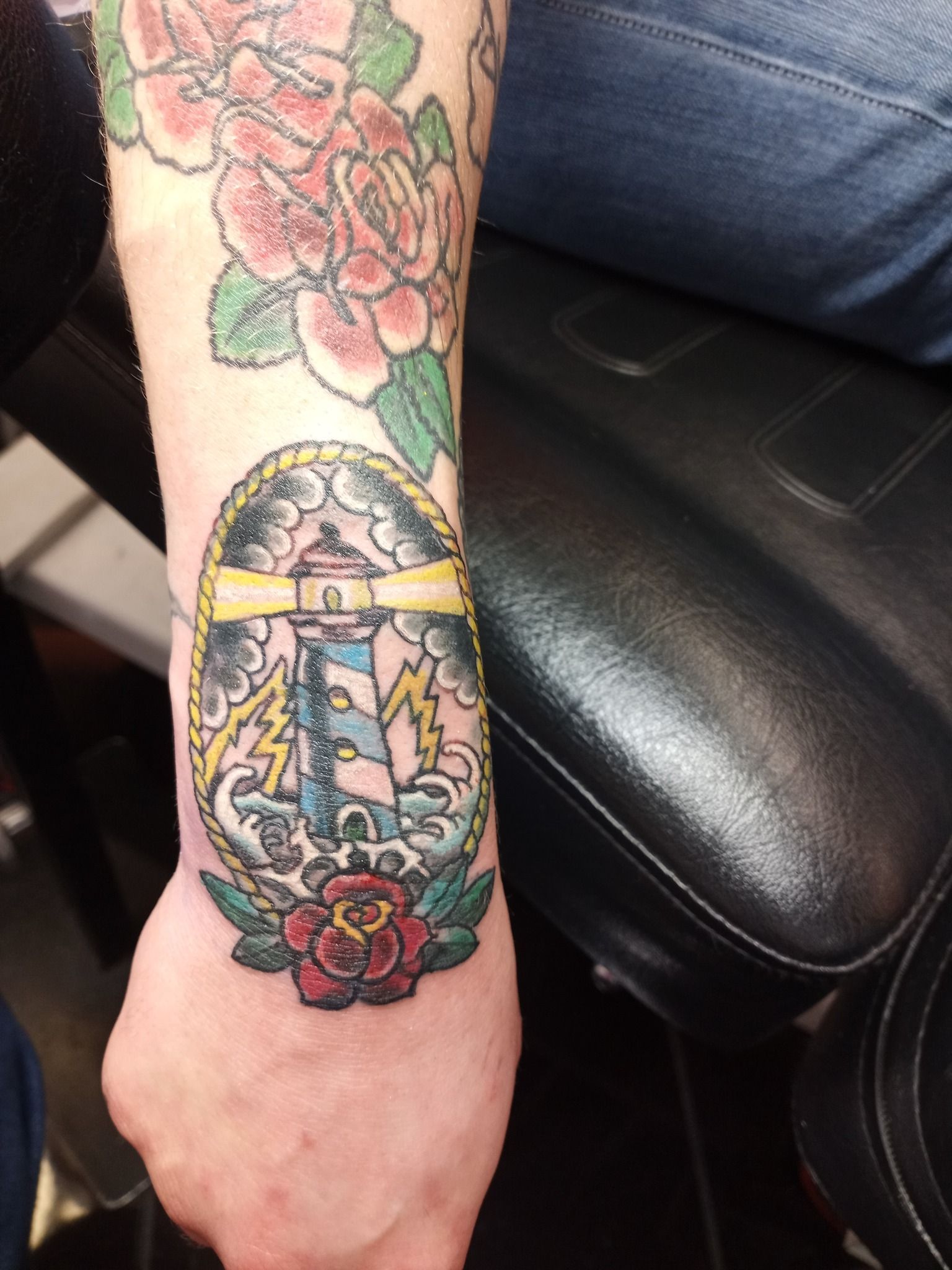 Tattoo of a lighthouse with lightning and roses on an arm, black leather chair in background.