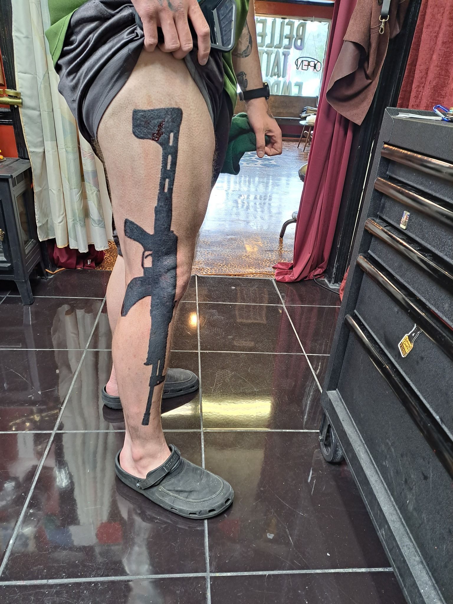 Person's leg with a black tattoo of an assault rifle; they stand in a shop wearing shorts and sandals.