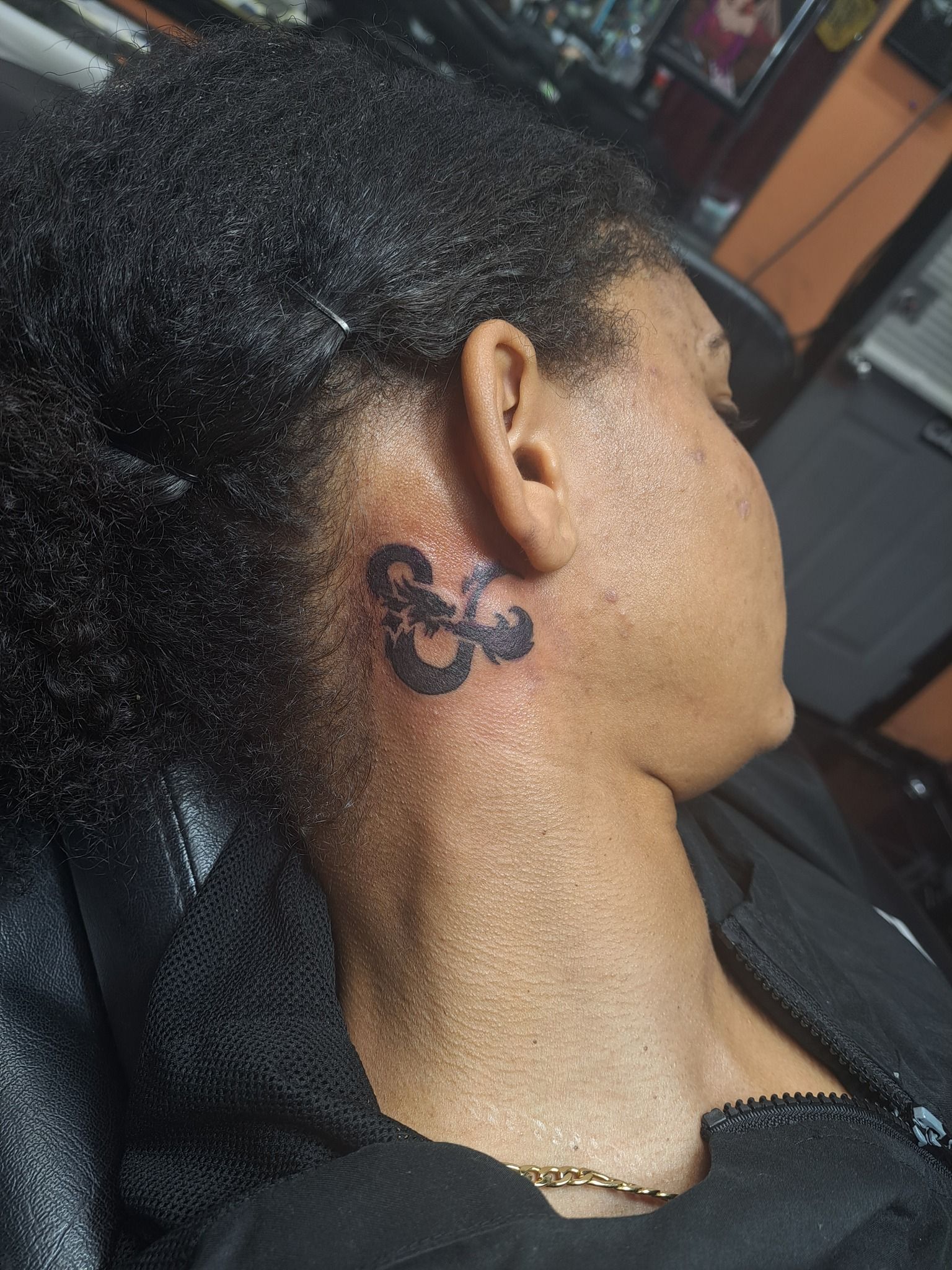 Black ink ampersand tattoo on a person's neck near the ear. Skin is tan. Black hair is pulled back.