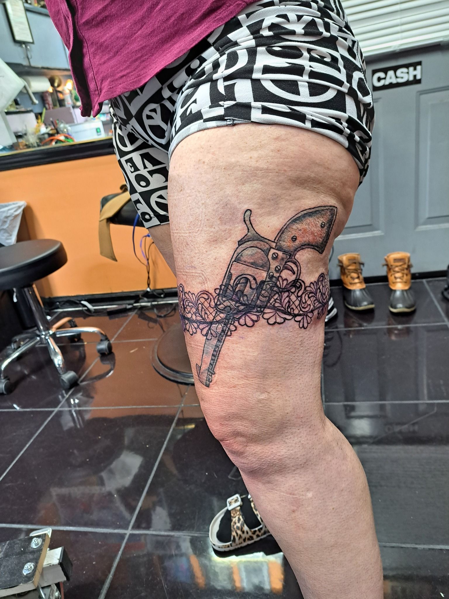 Leg with a black tattoo of a decorative garter and a horse head. Person wearing shorts, in a tattoo shop.
