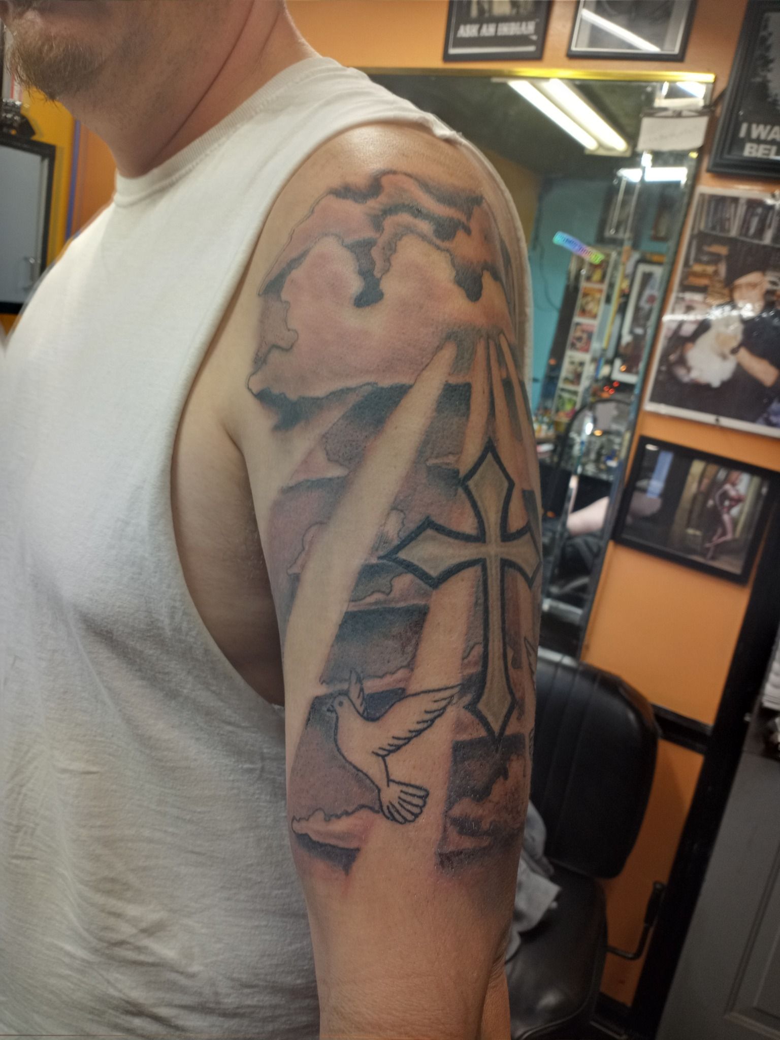 Tattoo on arm depicts a cross, dove, and clouds. Man wearing a white tank top. Tattoo studio background.