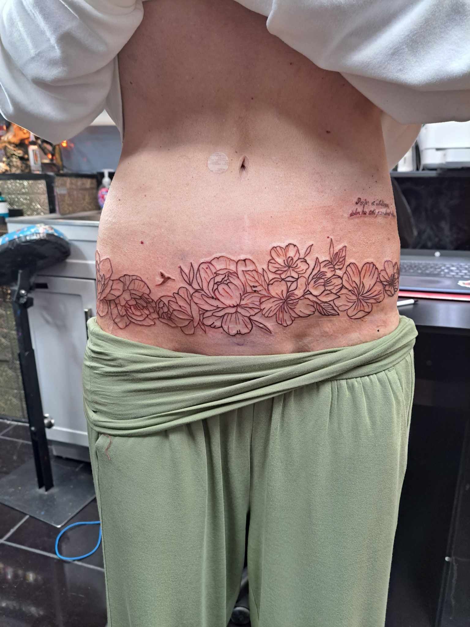 Tattoo of a floral design across a person's lower abdomen. The person wears green pants, a white shirt pulled up.