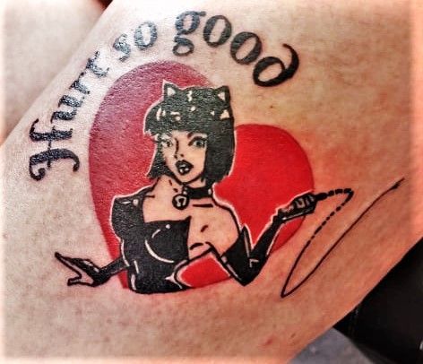Tattoo of a woman in black clothing and cat ears holding a whip, inside a red heart. Text reads "Hurt so good."