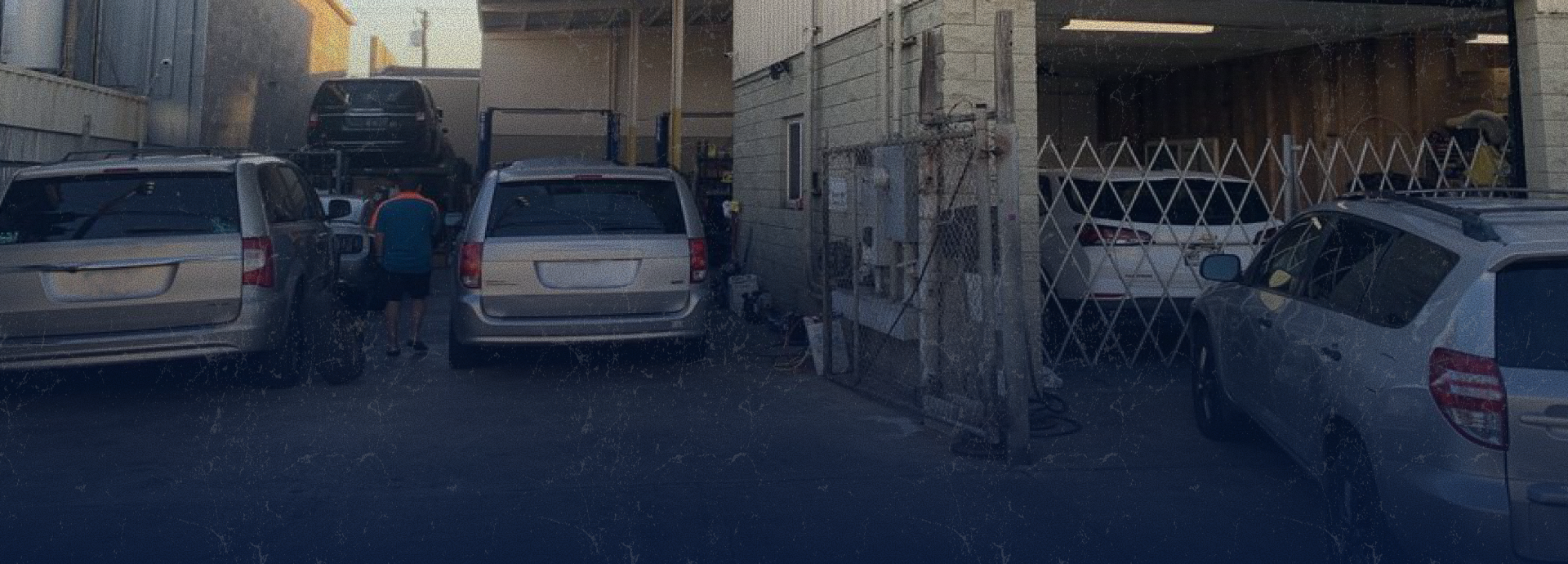 Cars parked in an open-air garage, person at work, metal gate, muted colors | Mike's Automotive AC