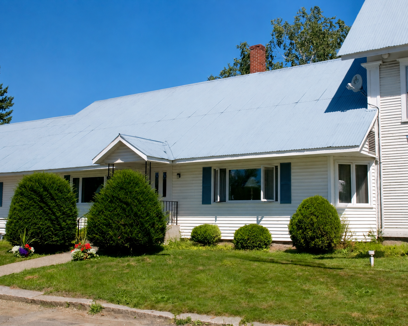White house with gray roof, trimmed shrubs, and a small front sign under a clear blue sky.
