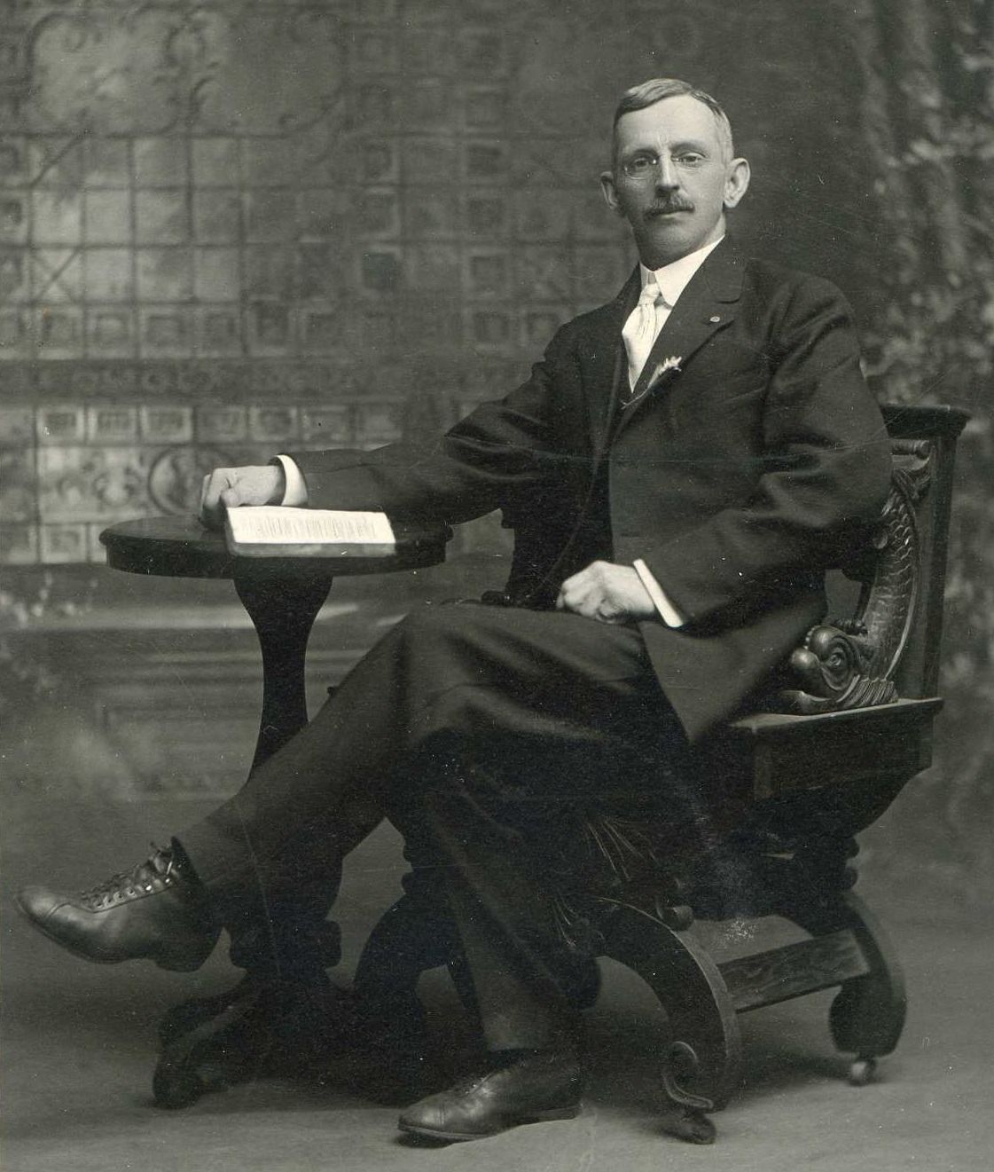 Man seated at a desk in a studio portrait, with a book and papers beside him.
