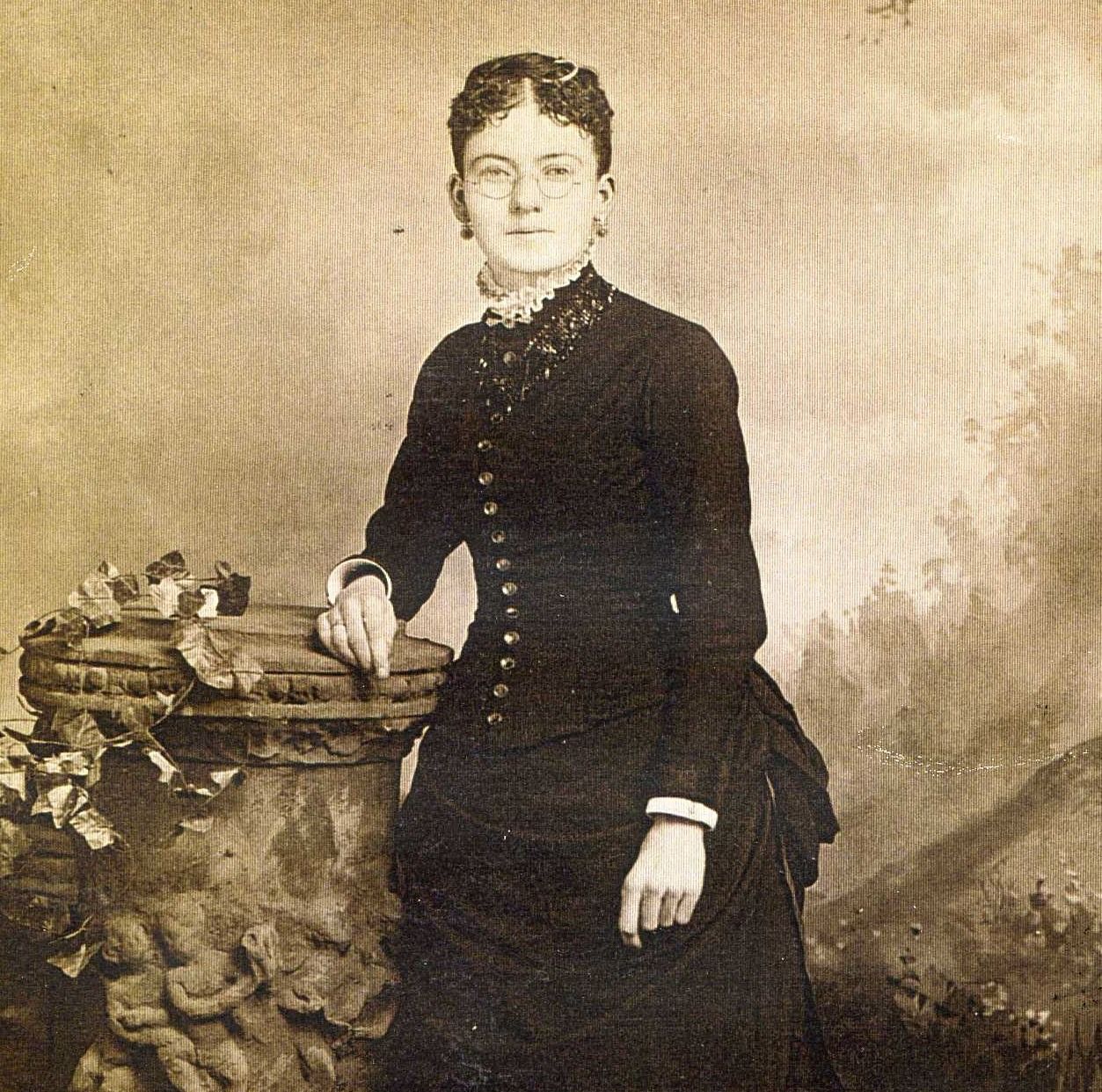 Sepia portrait of a woman in a dark dress standing beside a small table with flowers