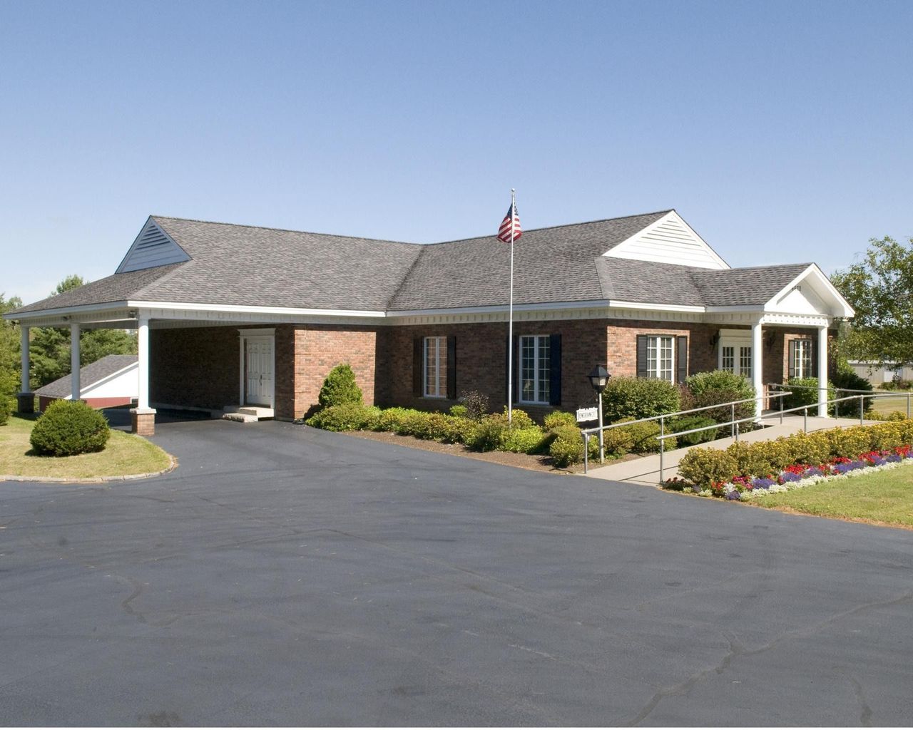 Brick building with white columns, a flagpole, and a paved parking lot under a clear blue sky