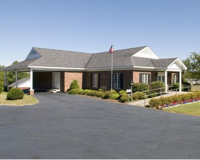 Brick building with white columns, a flagpole, and a paved parking lot under a clear blue sky