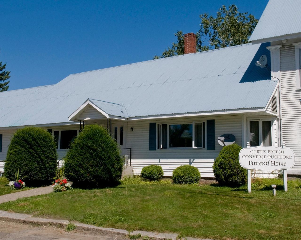 White house with gray roof, trimmed shrubs, and a small front sign under a clear blue sky.
