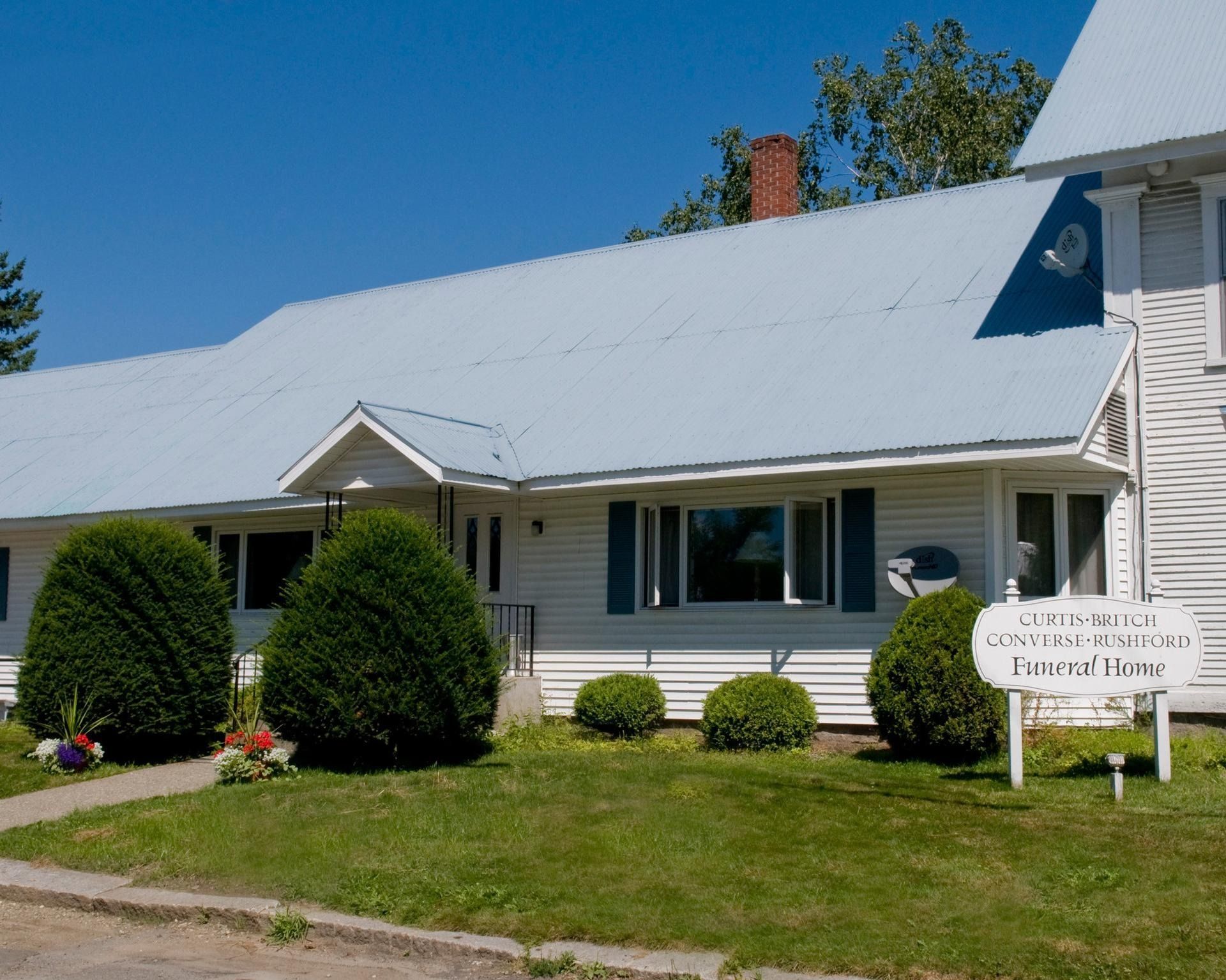 White house with gray roof, trimmed shrubs, and a small front sign under a clear blue sky.