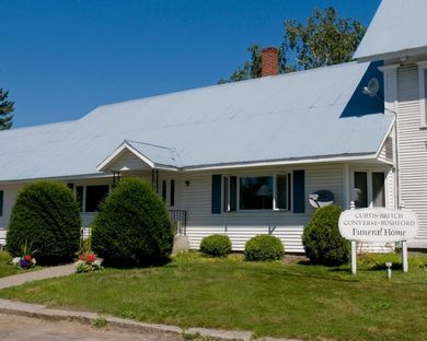 White house with gray roof, trimmed shrubs, and a small front sign under a clear blue sky.