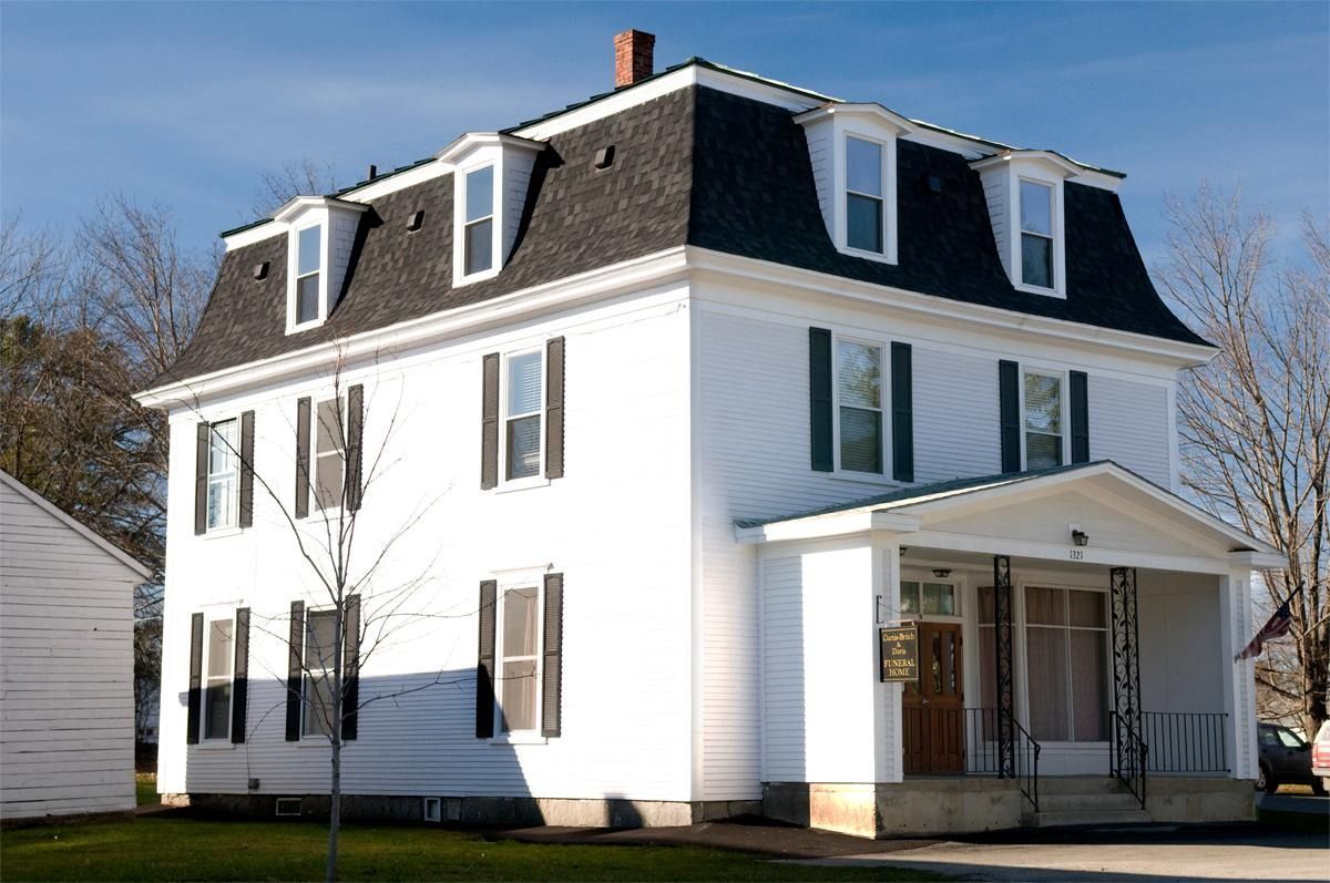 White two-story house with black mansard roof, front porch, and dormer windows on a sunny day