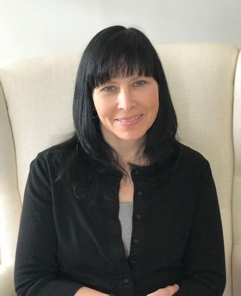 Person with black hair and bangs, wearing a black cardigan, sitting in a cream chair and smiling softly.