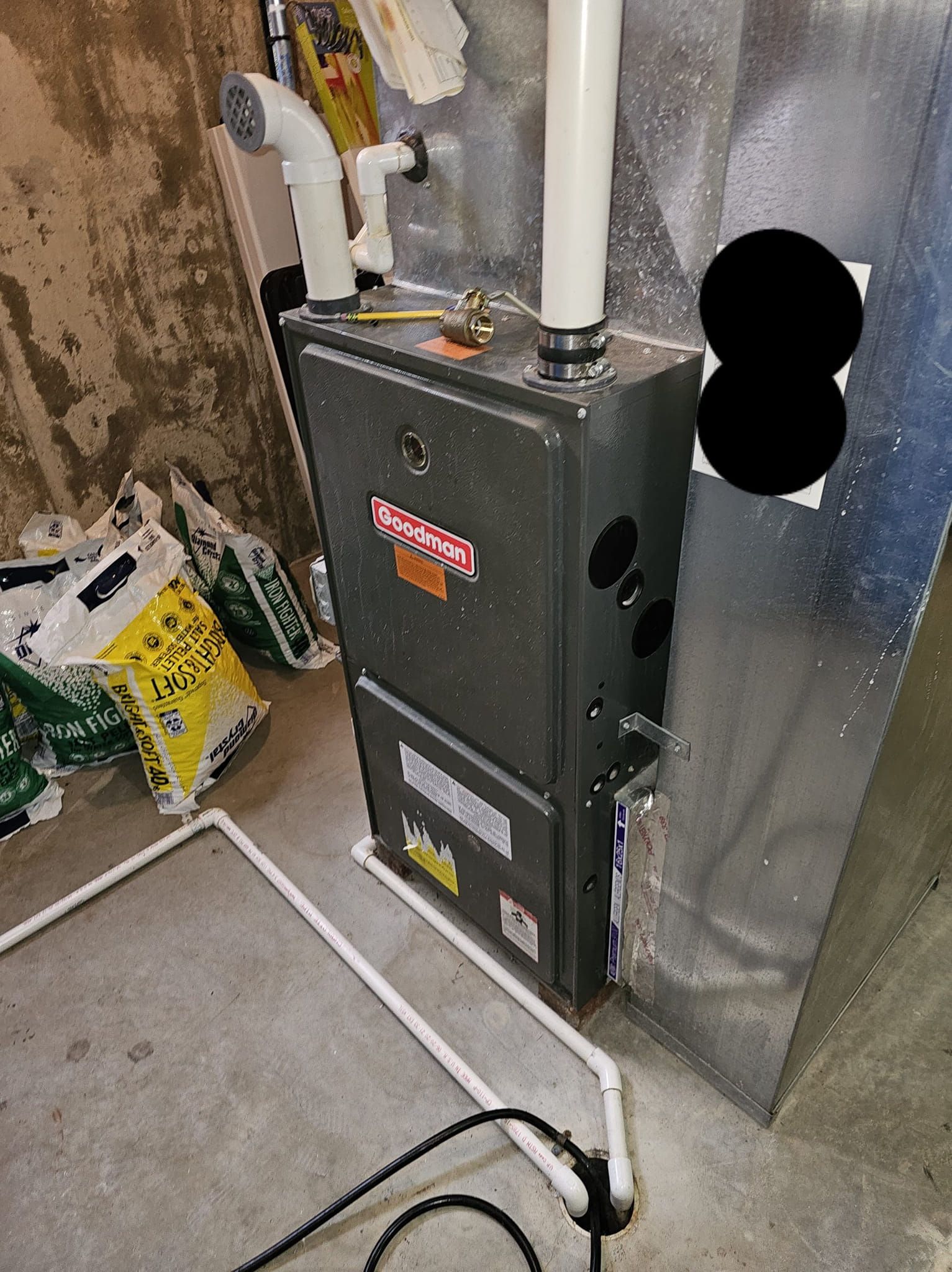 Goodman furnace and ductwork setup in a basement with PVC drain lines and nearby storage bags.