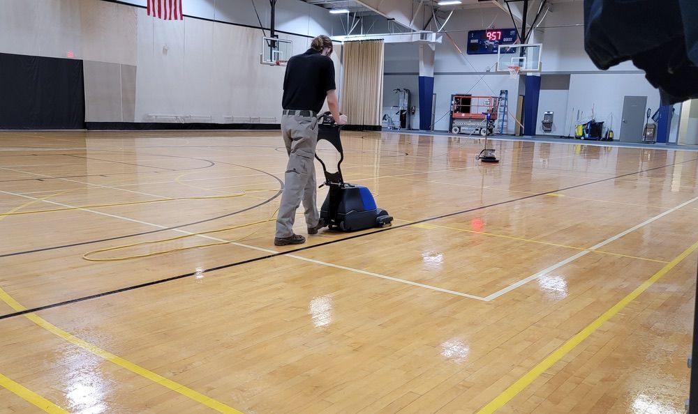 Technician performing commercial hardwood floor cleaning in a gymnasium with a floor scrubber machine.