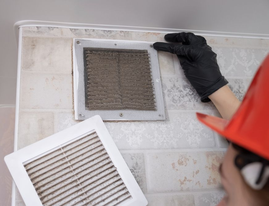 Person in black gloves cleaning a wall vent while holding a vent cover inside a tiled room.