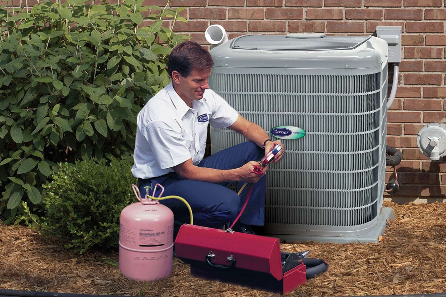 Technician cleaning an outdoor air conditioner unit beside a brick wall and garden bushes