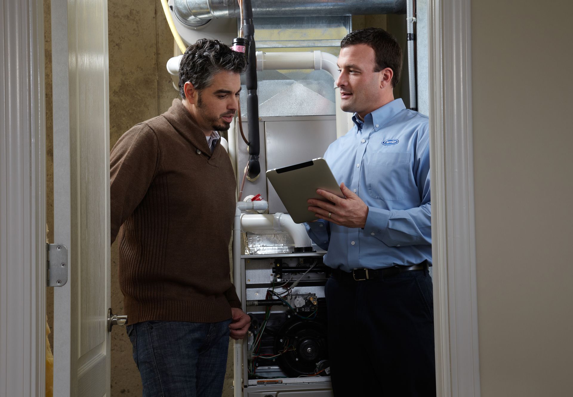 Two men talk in a utility room while one holds a clipboard.