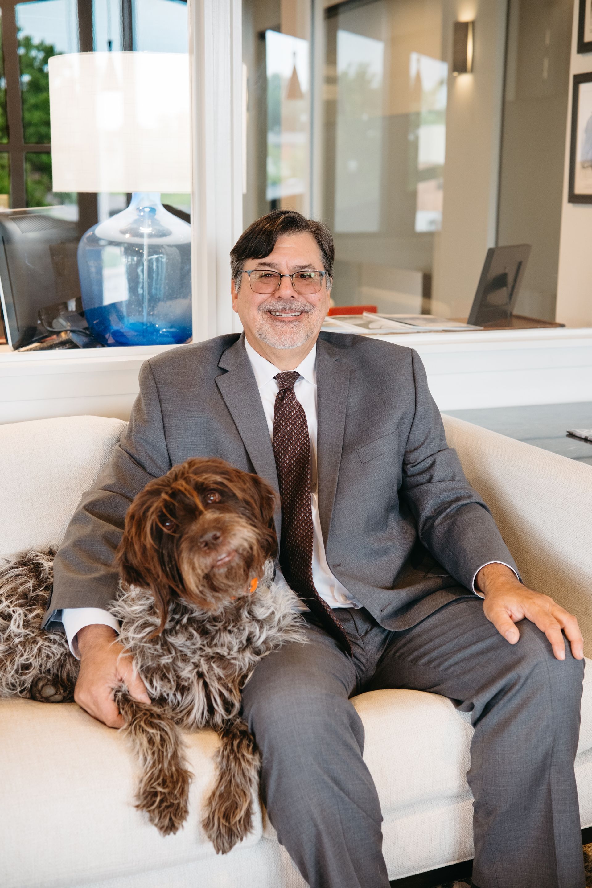 Patrick Bottaro wearing a gray suit, sitting on a beige couch with a brown dog.
