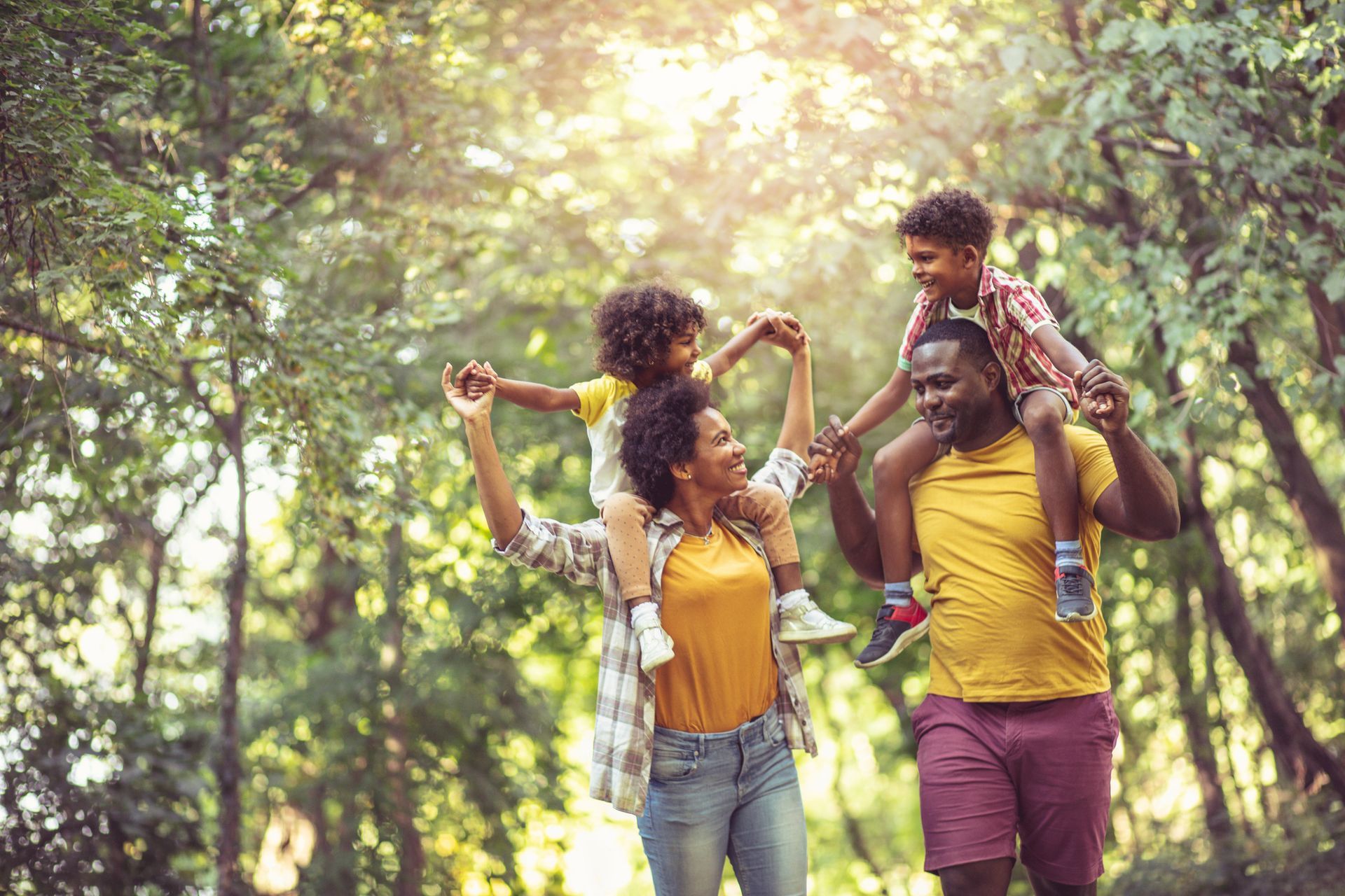 Two adults and a child walking through a sunlit forest, smiling and carrying the child on shoulders.