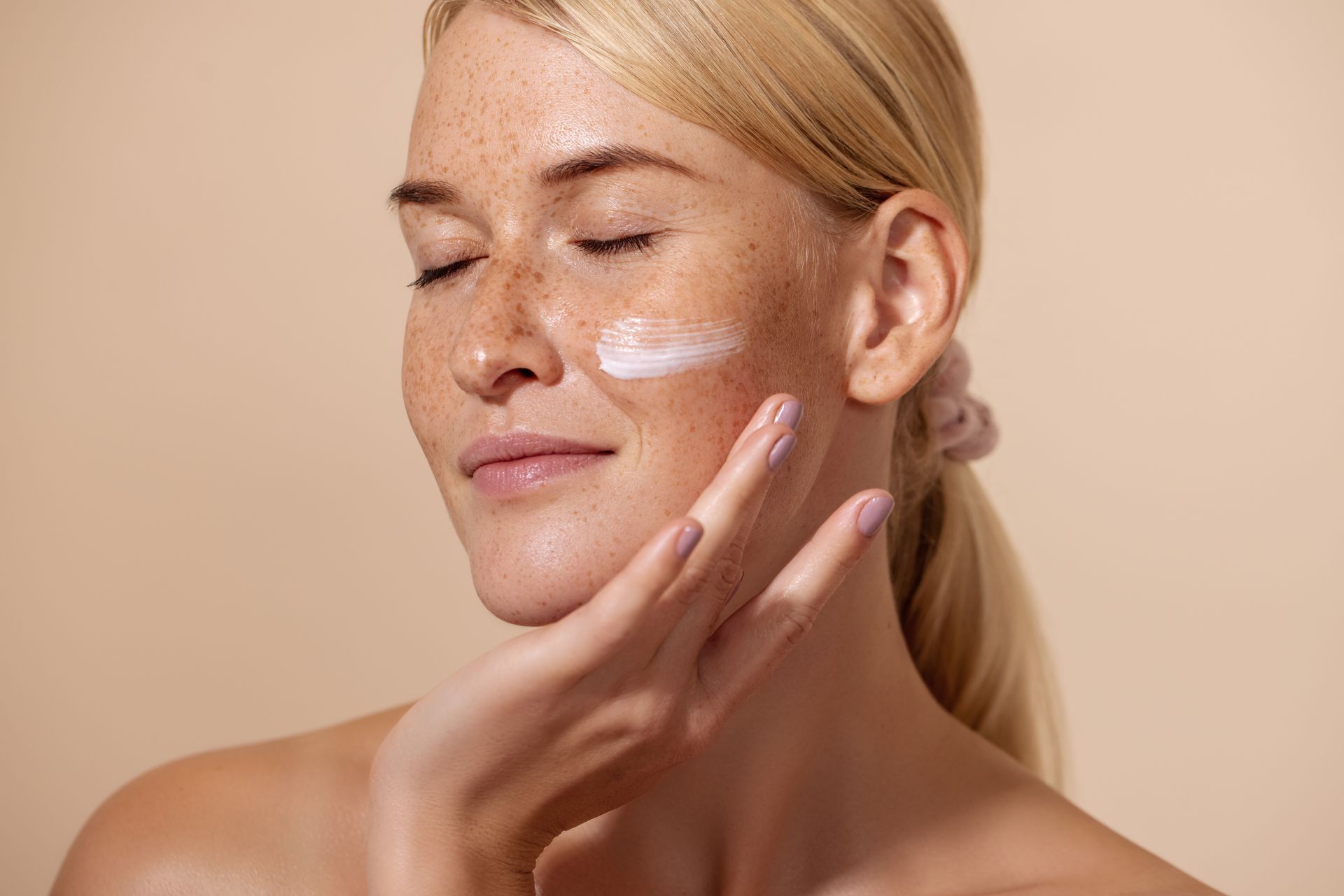 Woman applying skincare cream to her cheek with eyes closed and glowing skin
