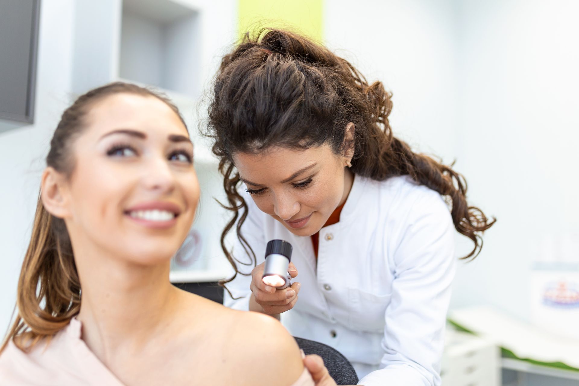 A dermatologist examining a smiling female patient's shoulder skin with a handheld dermatoscope. A dermatologist examining a smiling female patient's shoulder skin with a handheld dermatoscope.