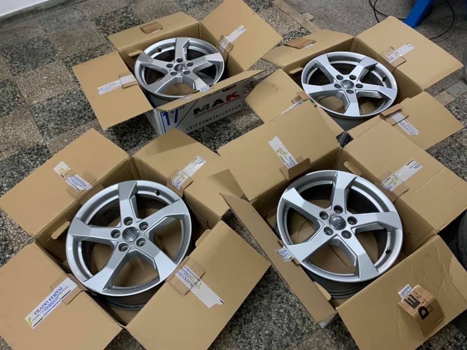 Cinque cerchi in lega d'argento per auto, in scatole di cartone aperte su un pavimento ricoperto di moquette.