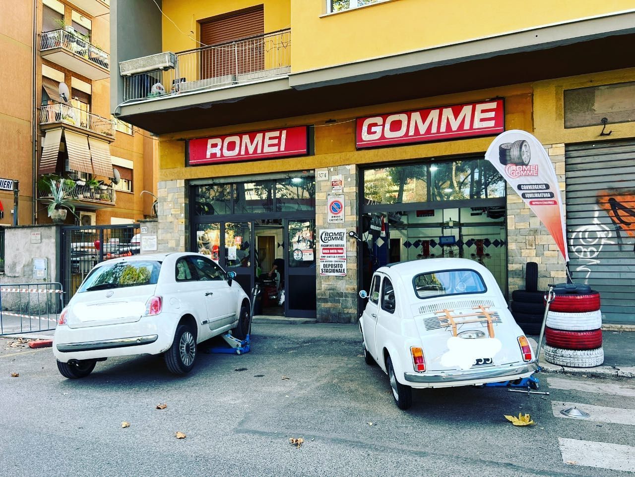 Vetrina di un'officina meccanica con due auto bianche parcheggiate all'esterno e insegne rosse con la scritta