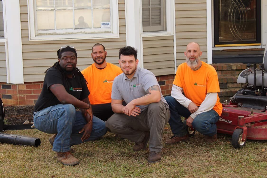 Four people wearing work shirts pose together in front of a house, with a lawn mower parked to their right.