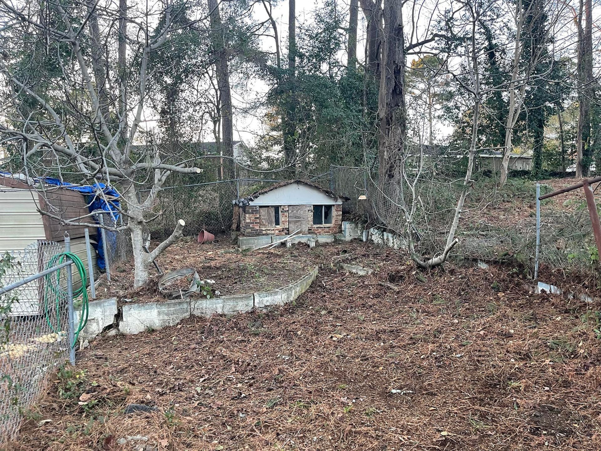 A small, brick-faced structure sits in a wooded backyard behind a chain-link fence and a concrete retaining wall.
