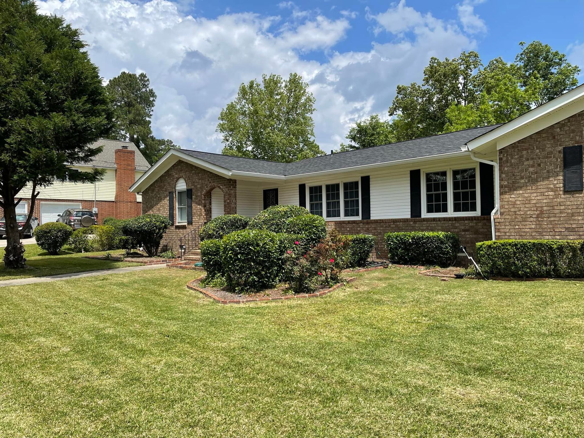 A single-story brick house with a dark roof and white trim sits behind a green lawn under a blue, cloudy sky.