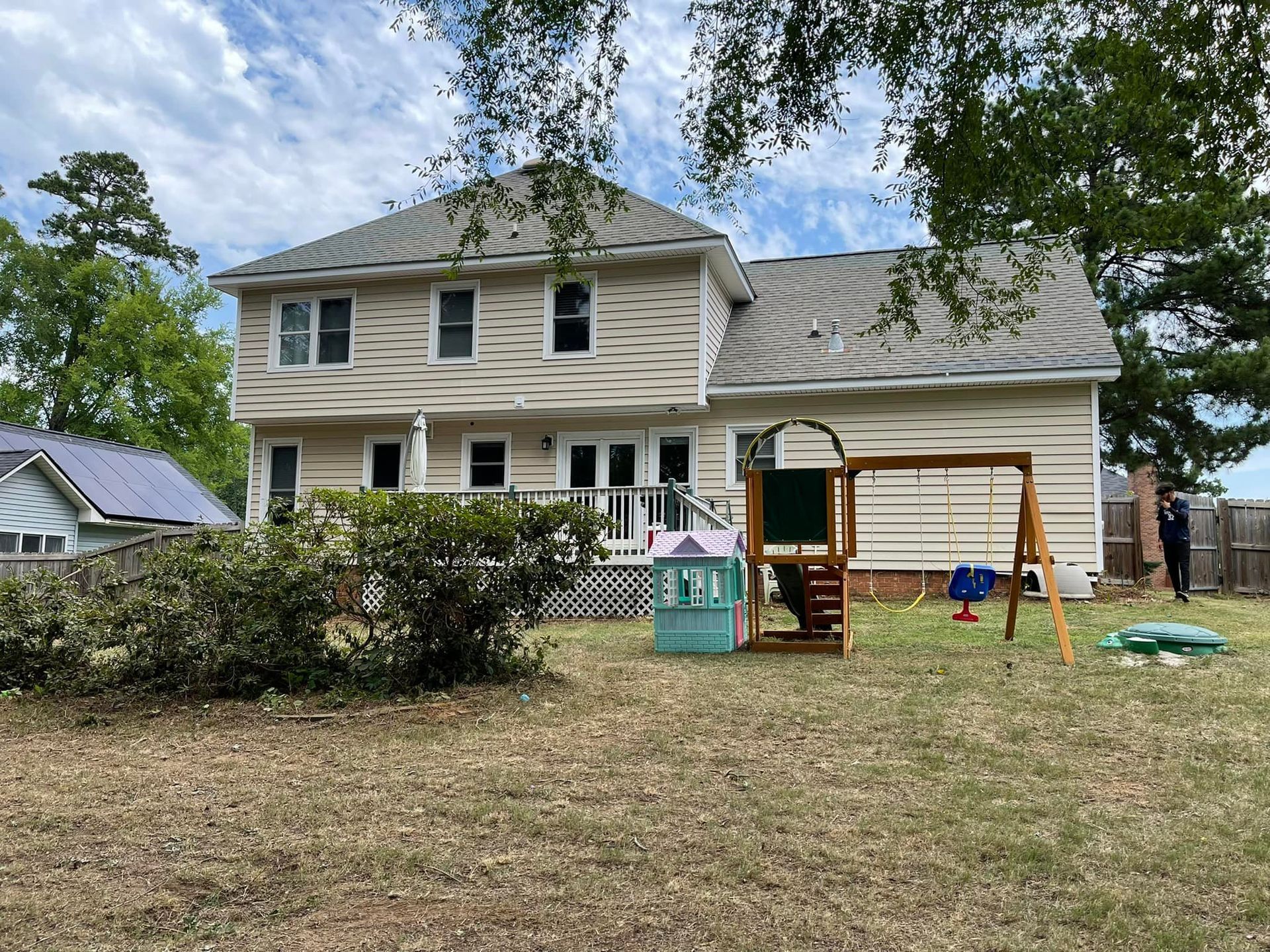 A two-story tan house with a wooden playset, small playhouse, and a large bush in the grassy backyard.