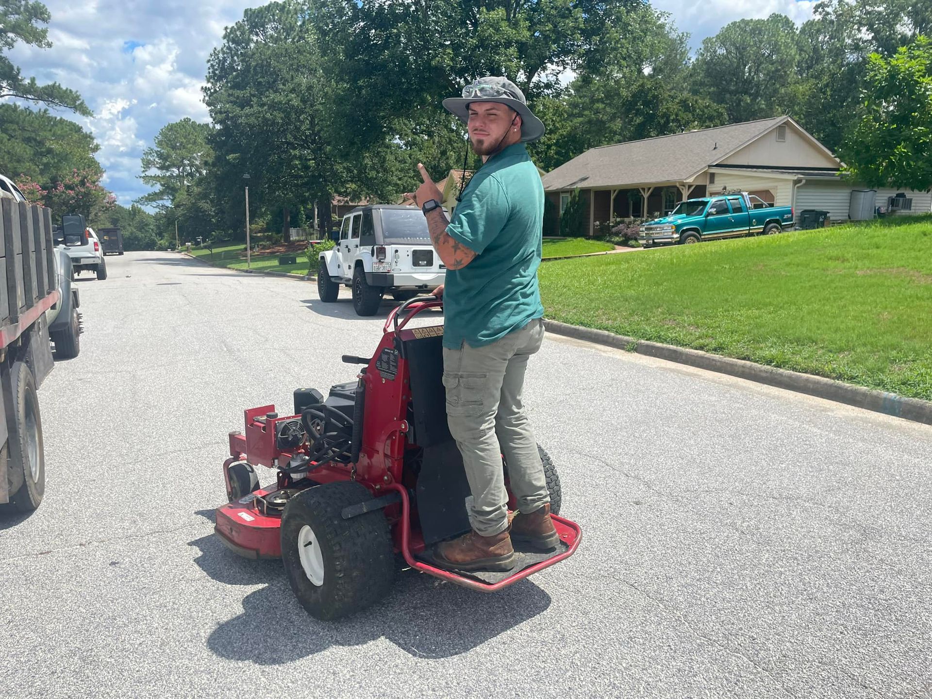 A person standing on a red stand-on lawn mower on a residential street, looking back and giving a thumbs up.