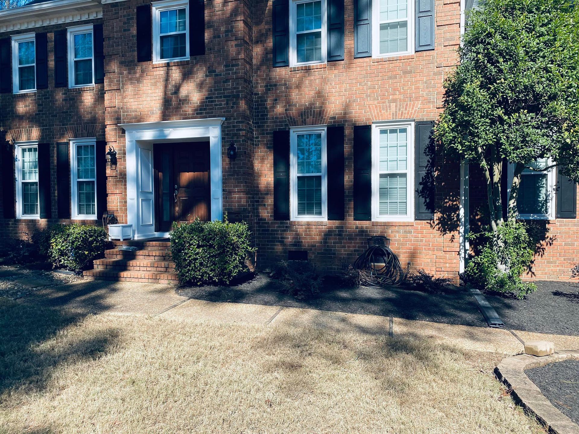 A two-story red brick house with black shutters, a white front entryway, and a tan yard with landscaping.