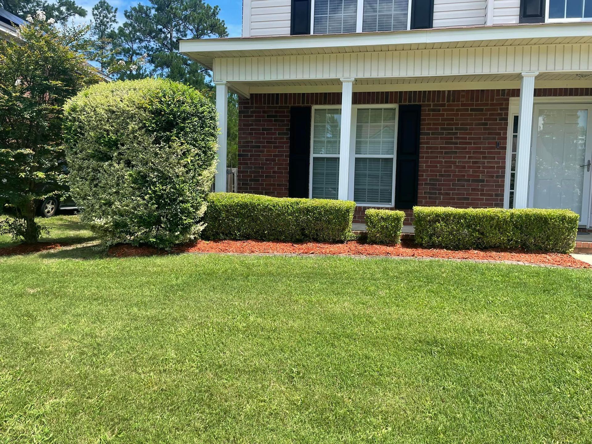 A brick house with a front porch, green lawn, a large rounded bush, and a low hedge along the foundation.