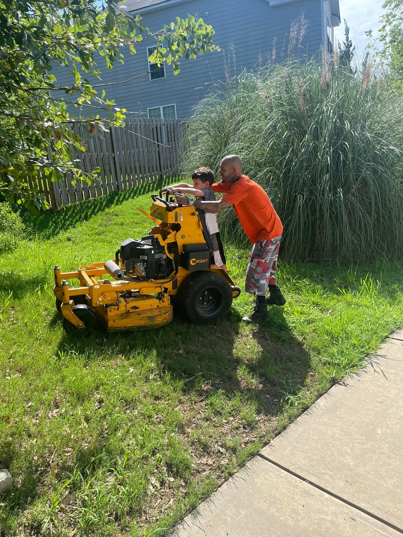 A person in an orange shirt helps a child stand on a yellow commercial lawn mower on a grassy lawn.